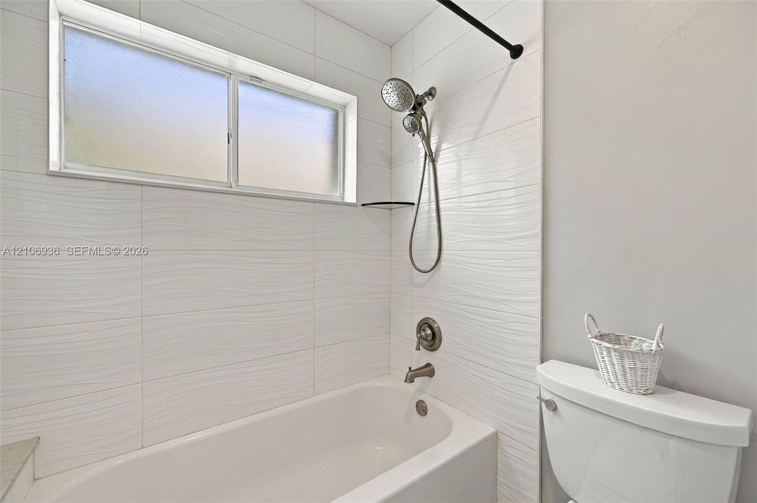 Bathtub featuring textured white wall tiling, a handheld showerhead fixture, and a horizontal sliding window