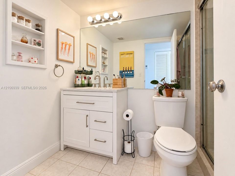 Bathroom featuring a vanity with shaker-style cabinetry, light-toned stone countertop, integrated shelving, large-format mirror, and glass-enclosed shower