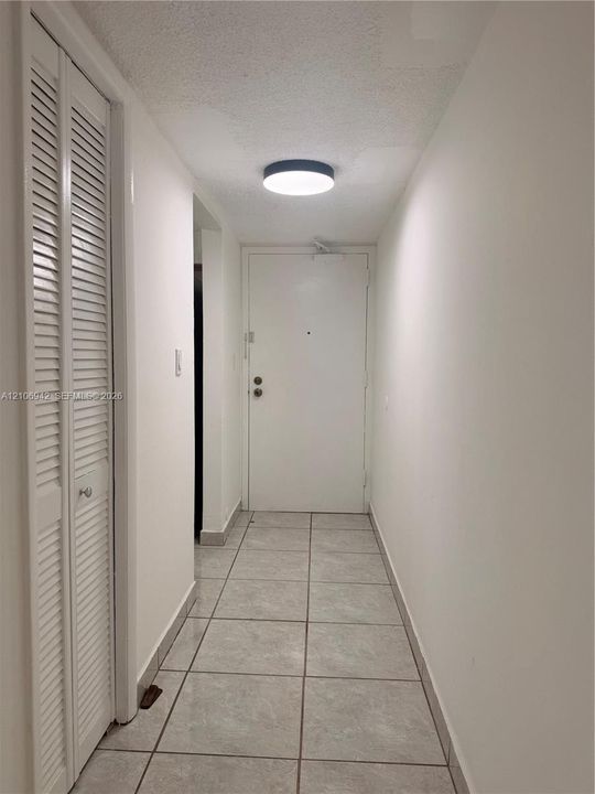Neutral interior hallway featuring large-format ceramic tile flooring, floor-to-ceiling bi-fold closet doors with louvered panels, and a flush-mount circular LED light fixture