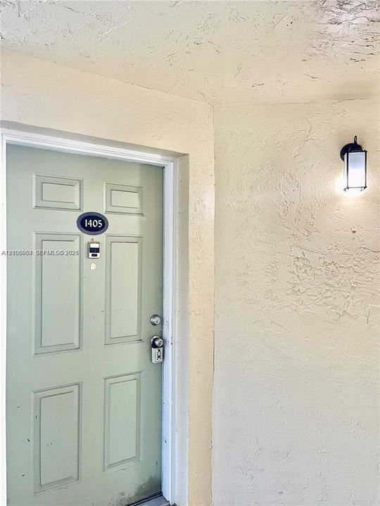 Paneled door with a metallic deadbolt, keyed entry lock, and rectangular wall-mounted exterior light fixture set against textured stucco walls