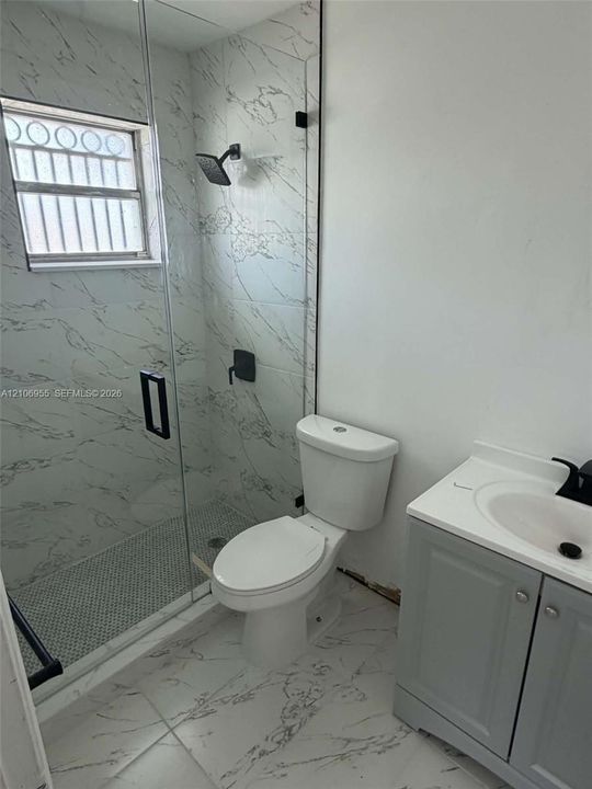 Walk-in shower featuring glass enclosure, wall-mounted matte black rainfall showerhead, and patterned tile surround, complemented by a grey vanity with white countertop and black fixtures