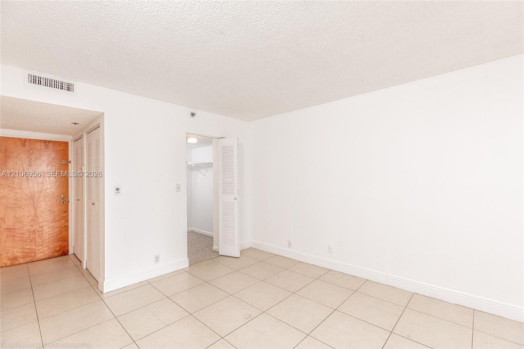 Interior space featuring neutral tile flooring, smooth-finish walls, a wooden entry door, and a built-in walk-in closet with louvered doors
