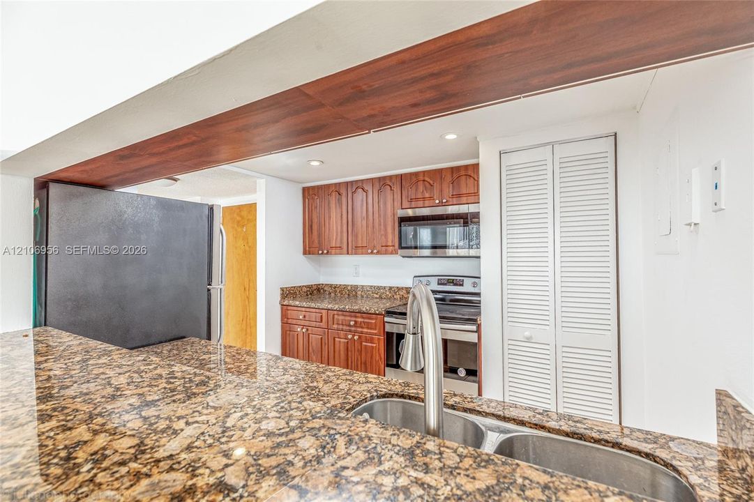Kitchen featuring stone countertops, wood-finish cabinetry, stainless steel double-basin sink with high-arc faucet, integrated microwave, and white louvered closet doors