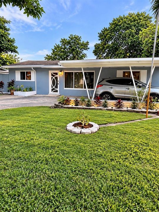Single-story exterior featuring a painted stucco finish, an attached angular carport, a decorative glass-panel front door, and a landscaped yard with stone garden borders