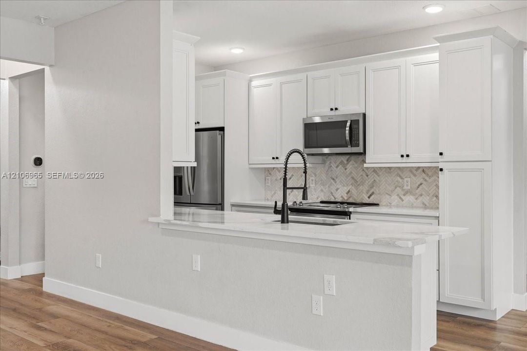 Kitchen featuring white shaker-style cabinetry, stone-finish countertops, herringbone-pattern tile backsplash, stainless steel appliances, and wood-finish flooring