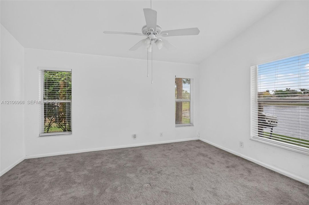 Neutral-toned interior featuring carpeted flooring, vaulted ceilings, a central ceiling fan with integrated lighting, and multiple windows with horizontal blinds