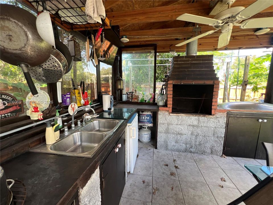 Outdoor kitchen featuring a built-in brick grill, tile flooring, dual-basin stainless steel sink, solid surface countertops, and an overhead ceiling fan
