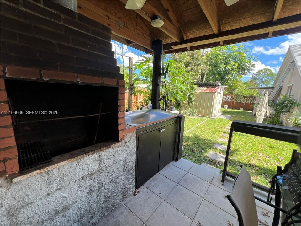 Covered outdoor kitchen featuring a built-in masonry grill, tiled flooring, exposed wood ceiling beams, and integrated cabinetry