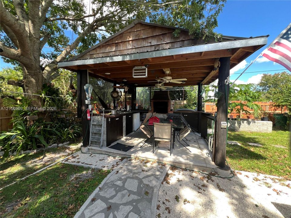 Covered outdoor kitchen pavilion featuring a gable roof, built-in cabinetry, integrated brick oven, and ceiling fans, set over a stone-paved patio