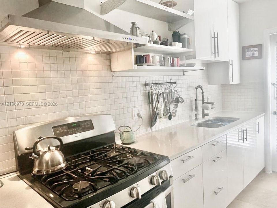 Kitchen featuring a stainless steel gas range, professional-grade range hood, white flat-panel cabinetry, white square-tile backsplash, and solid-surface countertops with a double-basin undermount sink