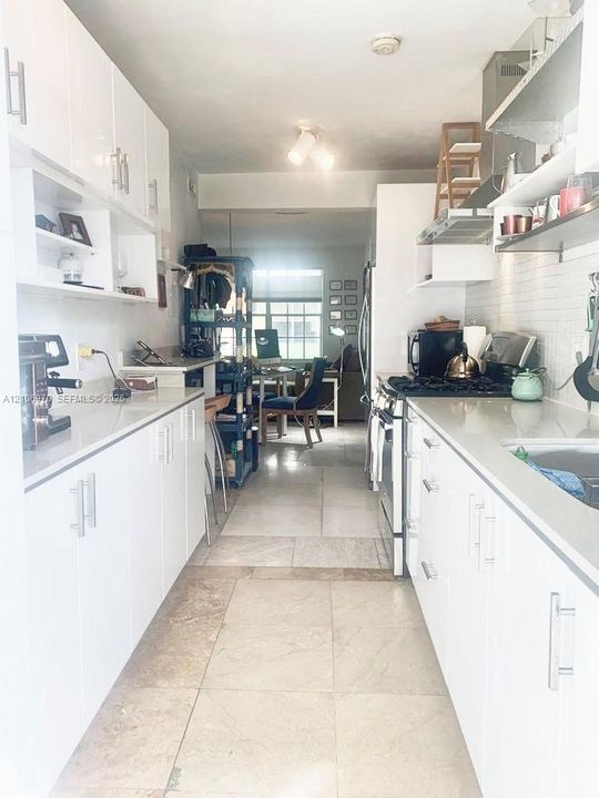 Galley kitchen featuring white flat-panel cabinetry, light-toned stone-finish flooring, solid-surface countertops, and a subway-style tile backsplash