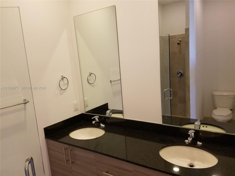Dual-sink vanity featuring wood-finish cabinetry, dark stone-style countertops, under-mount sinks, and chrome fixtures, paired with a glass-enclosed shower and wall-mounted mirror