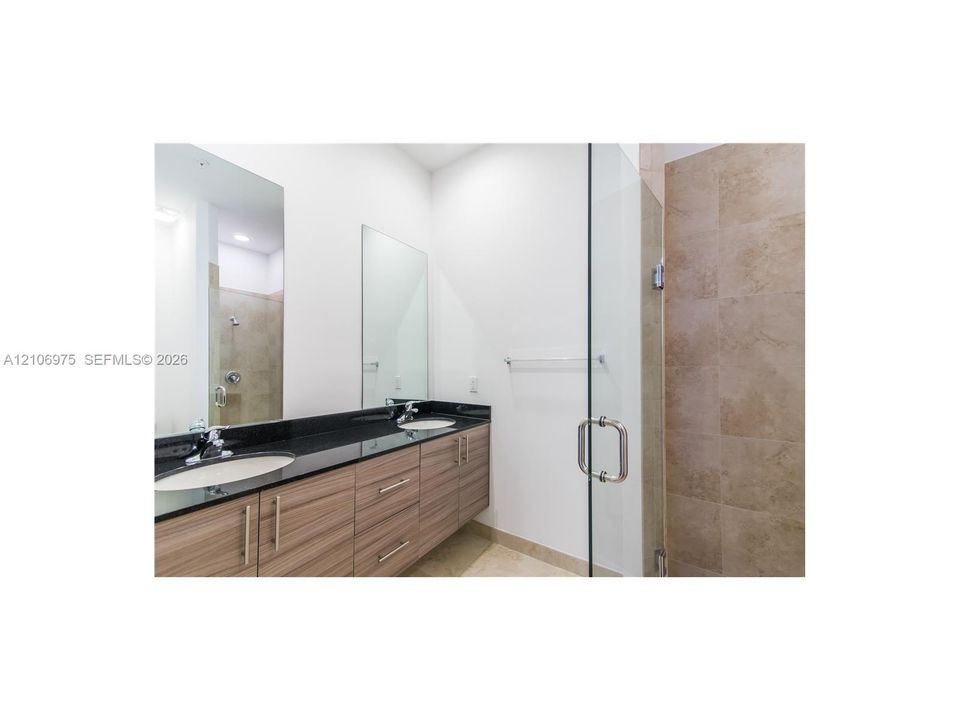 Floating wood-finish vanity with dual undermount sinks, black stone countertop, expansive wall-mounted mirror, and glass-enclosed shower with tiled surround
