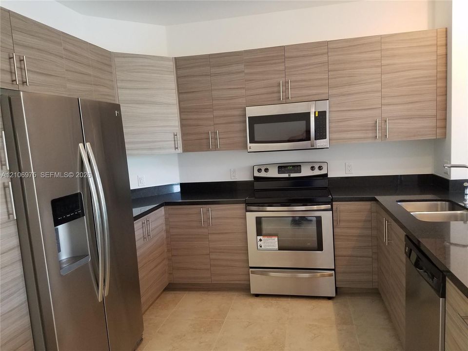 Kitchen featuring wood-finish cabinetry, stainless steel appliances, dark stone-surface countertops, dual-basin stainless steel sink, and tile flooring