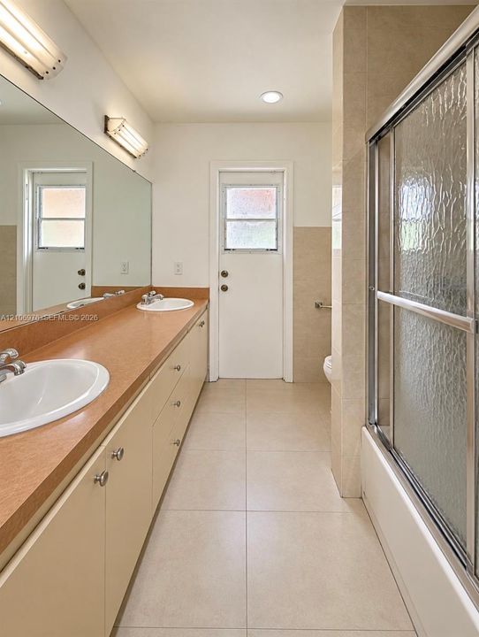Extended vanity with dual undermount sinks, solid-surface countertop, full-width wall mirror, and tub-shower combination with sliding textured glass doors