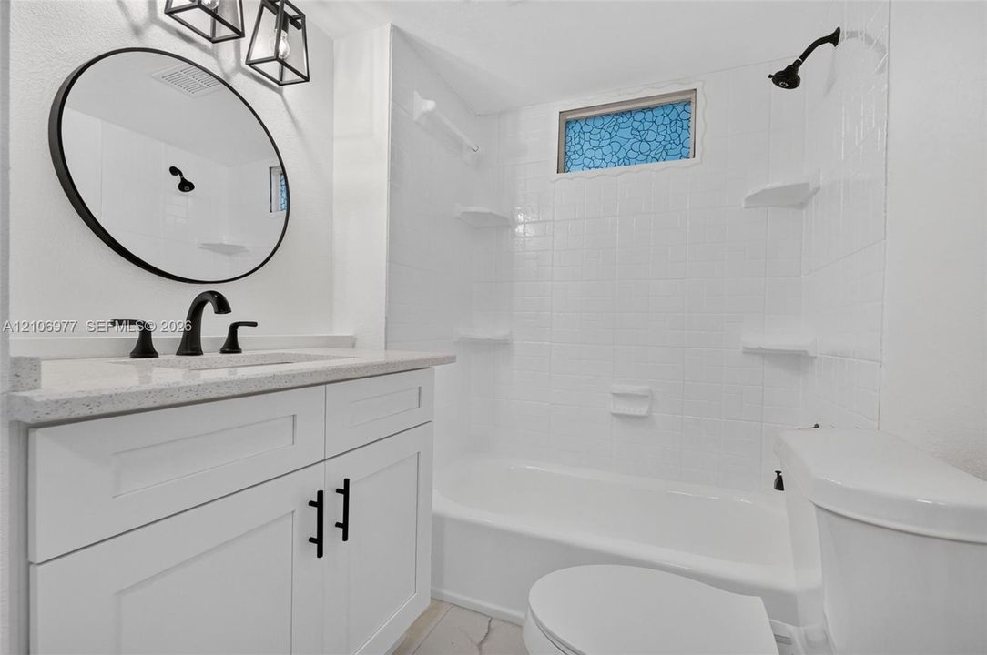 Bathroom featuring a white shaker-style vanity with stone-composite countertop, circular vanity mirror, matte black plumbing fixtures, and a tiled shower surround with integrated shelving
