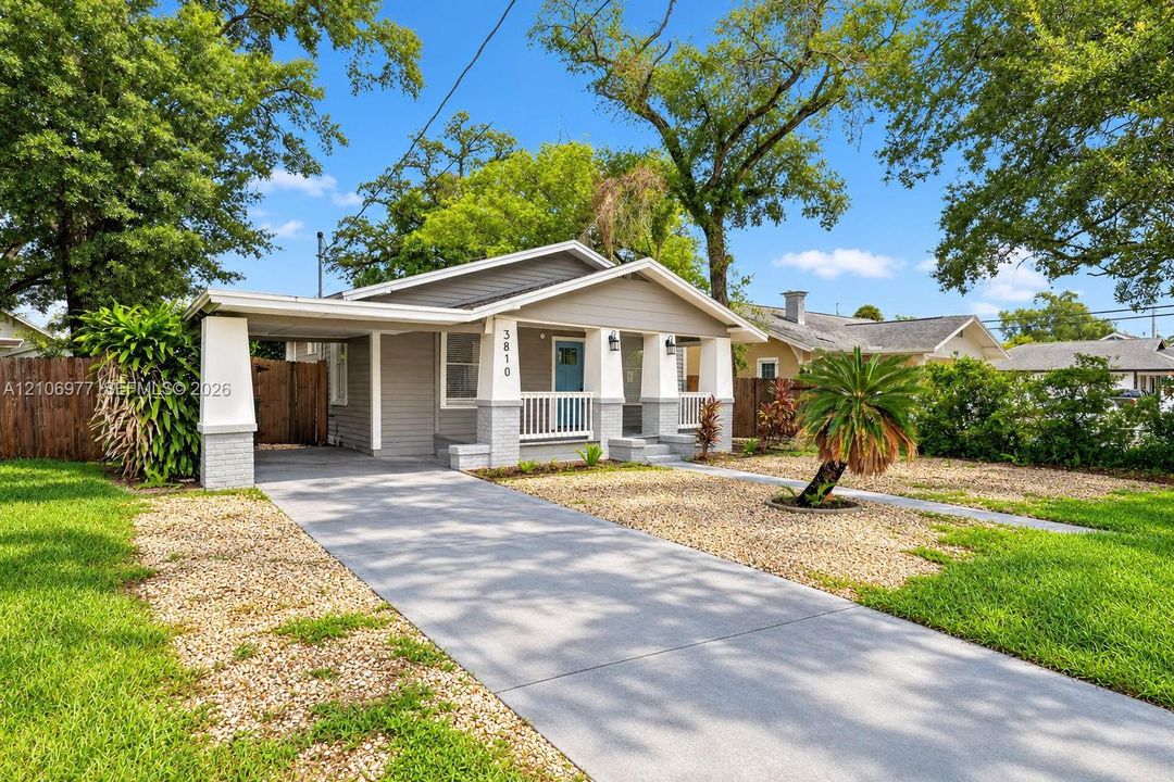 Single-story exterior featuring a covered front porch with tapered columns, gray horizontal siding, attached carport, concrete driveway, and xeriscaped front grounds