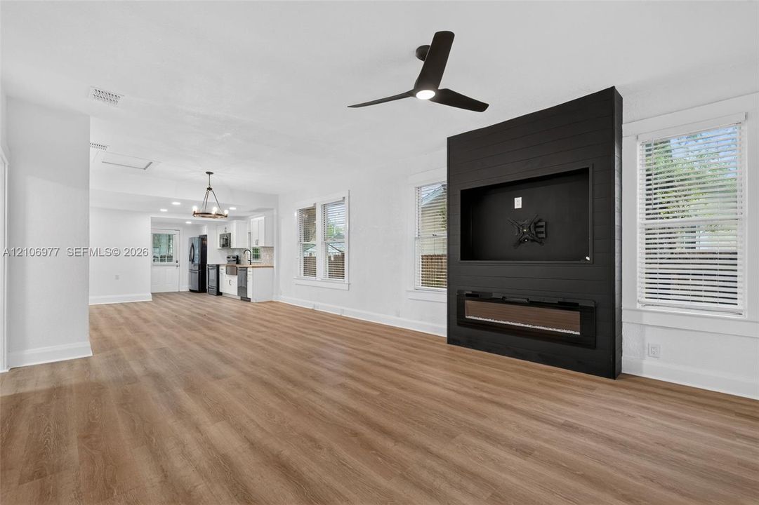 Open-concept space featuring wood-finish flooring, a matte black shiplap accent wall with a built-in electric fireplace, and a three-blade ceiling fan