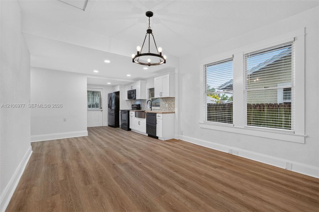 Open-concept living space featuring wood-finish flooring, white cabinetry, a farmhouse-style apron sink, matte black fixtures, and a statement chandelier