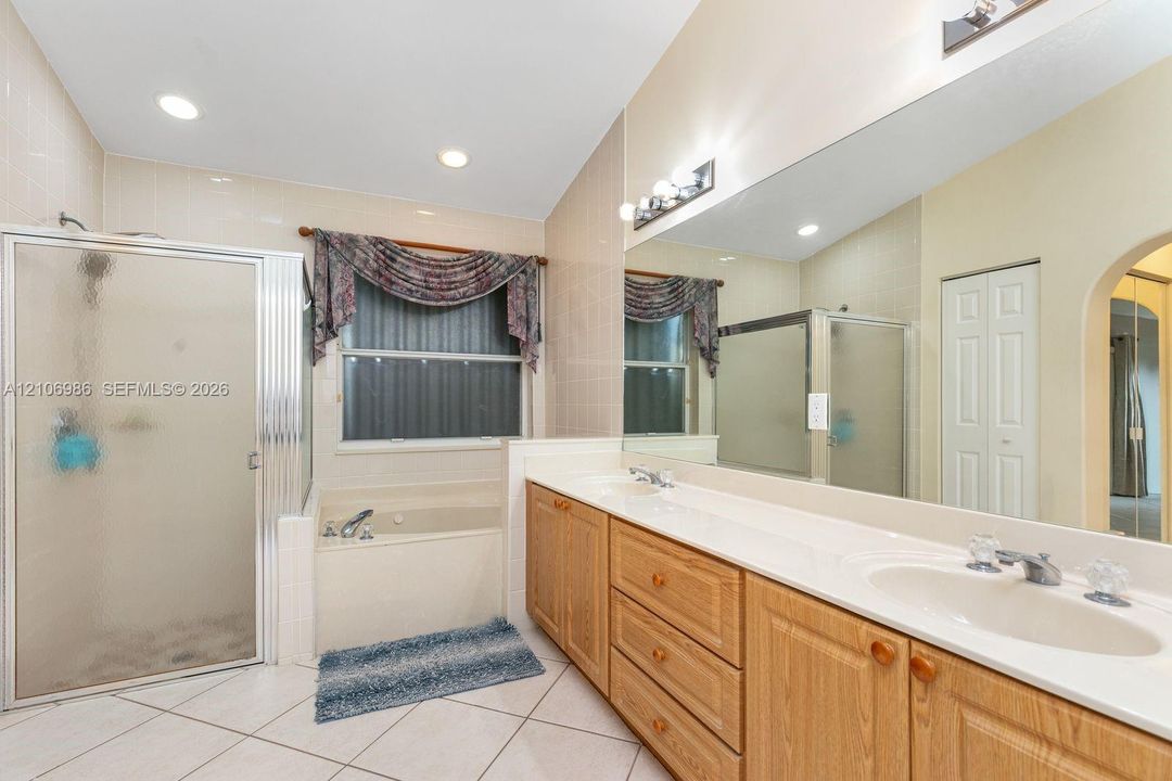 Bathroom featuring a dual-sink vanity with light wood cabinetry, a tiled-in soaking tub, a separate glass-enclosed shower, a full-width wall mirror, and tile flooring