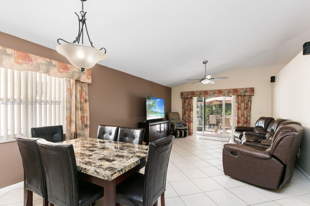 Open-concept area featuring light-toned tile flooring, vaulted ceilings, a central ceiling fan with integrated lighting, and glass sliding doors providing exterior access