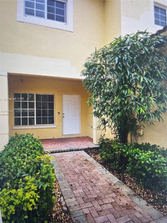 Exterior facade featuring a covered entryway, paved brick walkway, grid-patterned windows, and mature landscaping