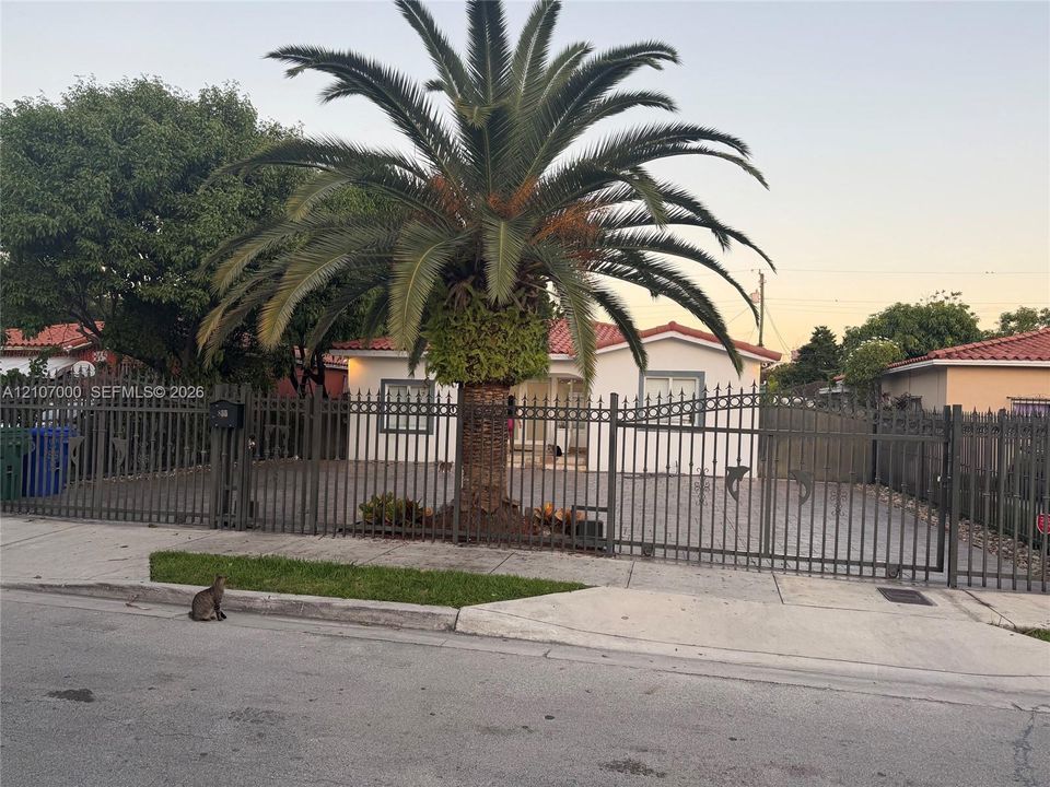 Stucco exterior featuring a terracotta tile roof, gated perimeter fencing, and a mature palm tree