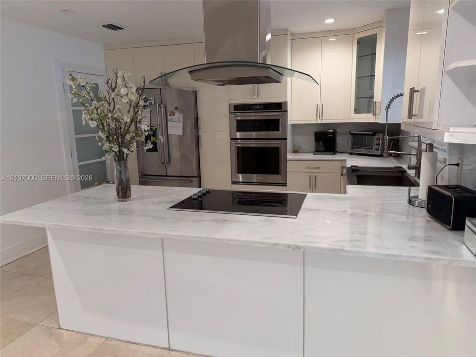 Kitchen featuring a stone-surface island with an integrated cooktop, stainless steel double wall ovens, a glass and stainless steel vent hood, and high-gloss cabinetry