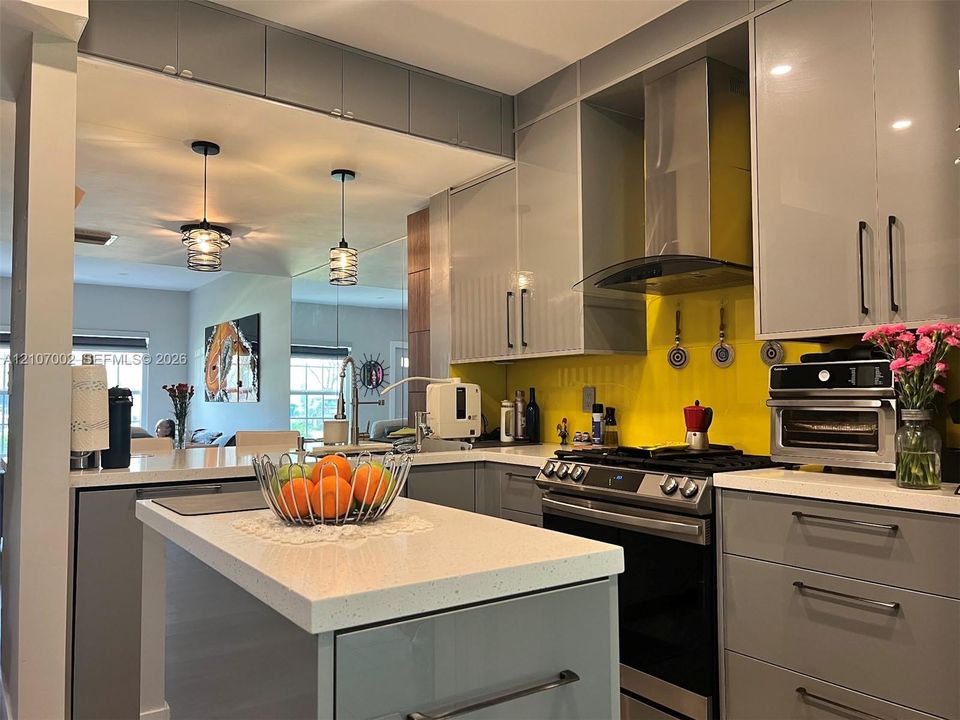 Contemporary kitchen featuring flat-panel cabinetry, a solid-surface island countertop, stainless steel range with overhead vent hood, and dual glass pendant lighting