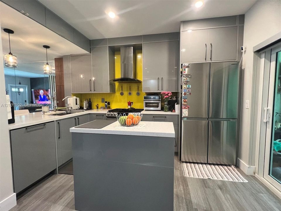 Kitchen featuring grey flat-panel cabinetry, a central island with a solid-surface countertop, stainless steel refrigerator, vibrant backsplash, and recessed lighting