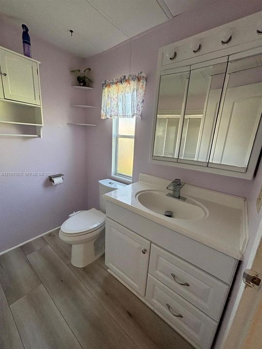 White vanity featuring integrated cabinetry, a composite sink, and a single-handle faucet, complemented by a tri-fold mirrored medicine cabinet, wood-finish flooring, and wall-mounted shelving