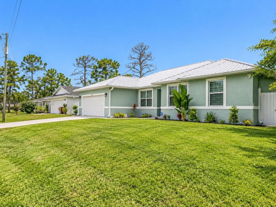 Single-story residence featuring a light green stucco exterior, white metal roof, attached two-car garage, concrete driveway, and landscaped front yard