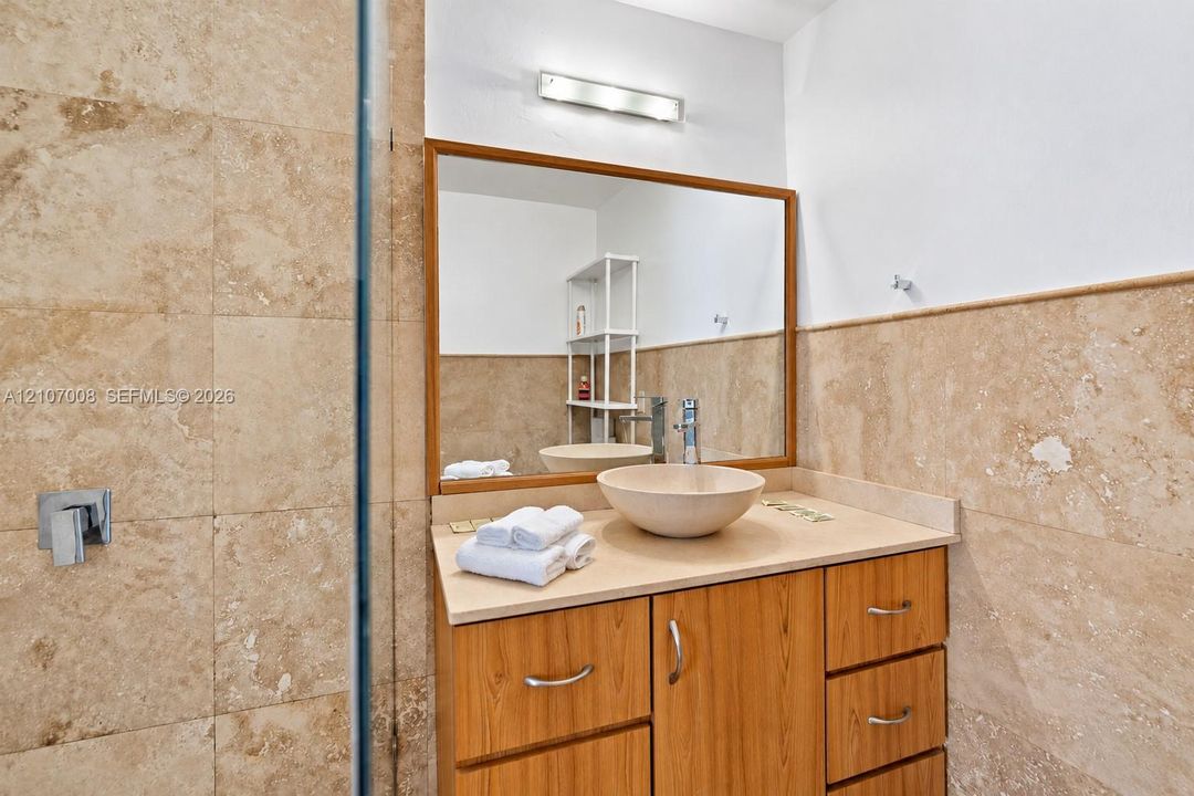 Vanity featuring wood-finish cabinetry, a stone-composite countertop, a circular vessel sink, a chrome single-handle faucet, and large-format neutral-toned tile wall cladding