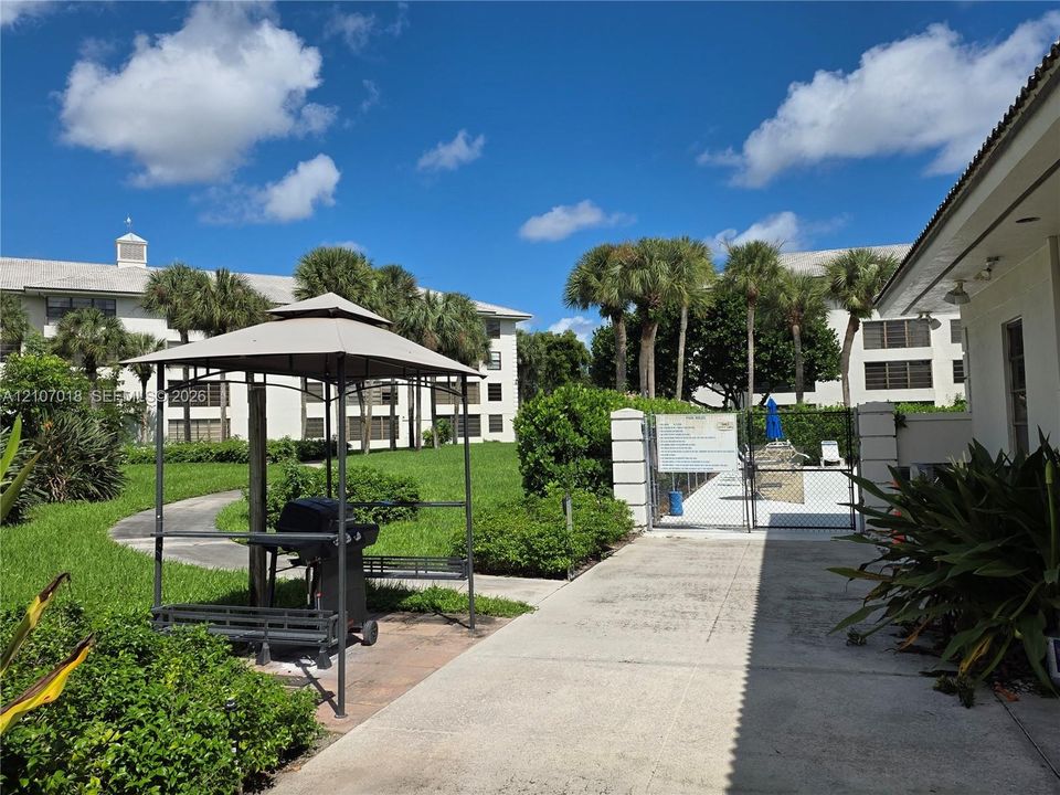 Outdoor common area featuring a paved walkway, manicured green space, mature palm trees, and a covered grilling station