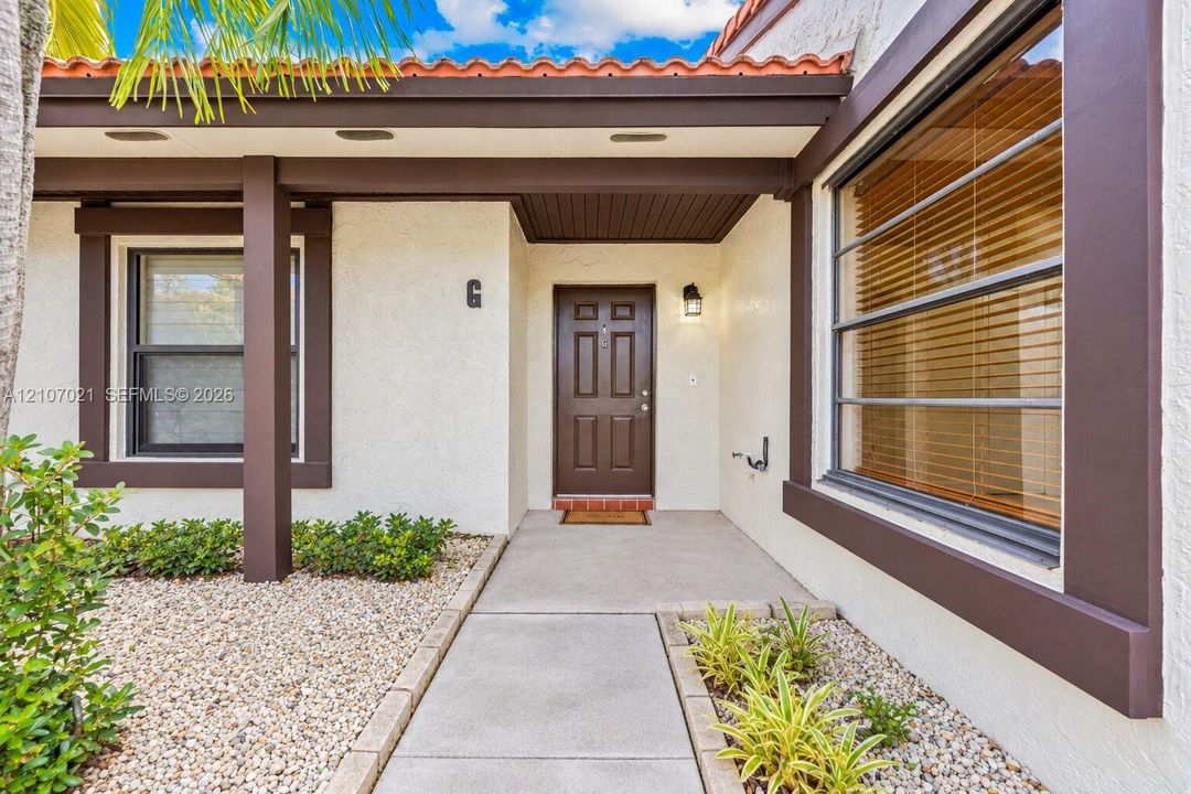 Covered entryway featuring textured stucco exterior, terra cotta roof tiles, solid-core panel door, horizontal window shutters, and low-maintenance rock landscaping