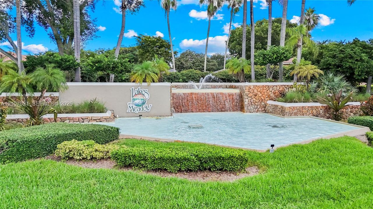 Landscaped entrance featuring a tiered rock waterfall, stone masonry walls, mature palm trees, and manicured shrubbery