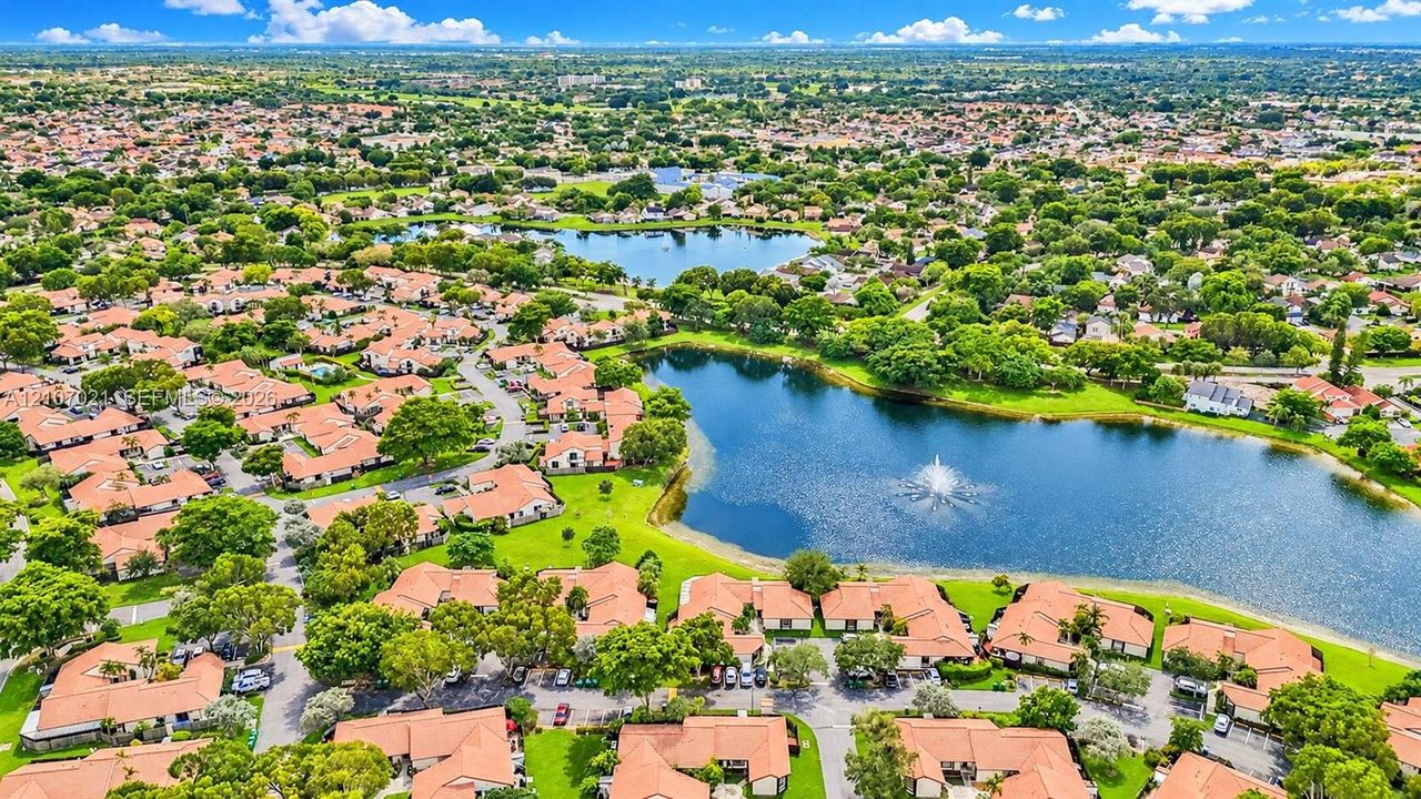 Aerial perspective featuring multi-unit structures with terracotta tile roofing, mature landscaping, and waterfront scenery including a central fountain