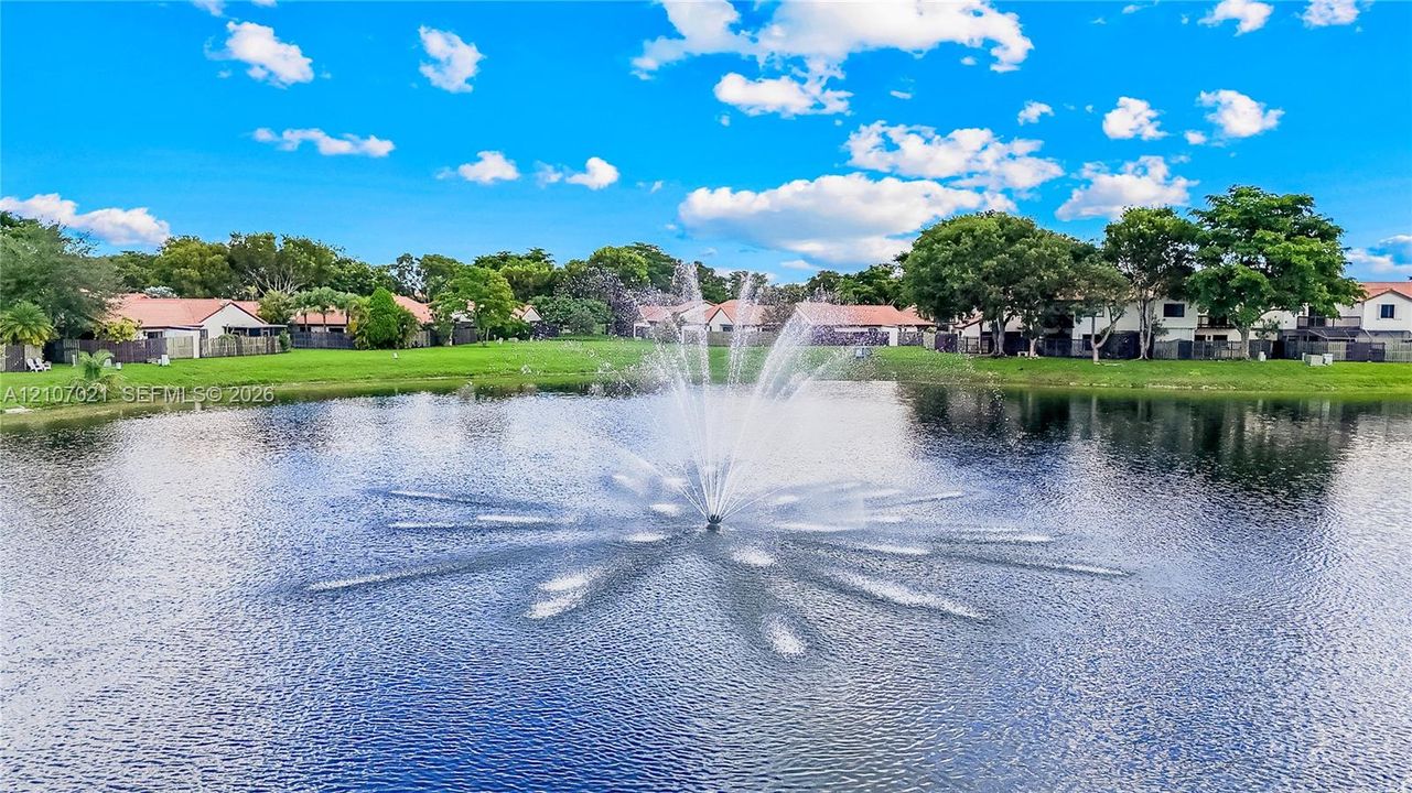 Water feature fountain positioned within a pond, surrounded by landscaped grass areas and mature trees