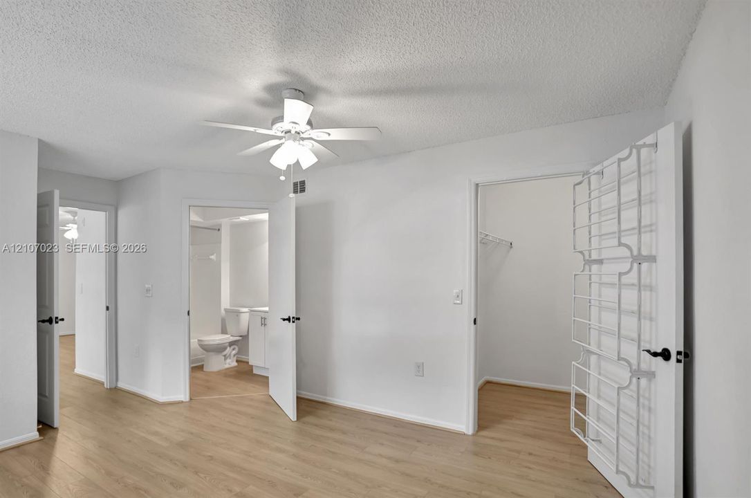 Room featuring light wood-finish flooring, a ceiling fan with integrated lighting, and multiple interior access points including a bathroom with a white vanity and a walk-in storage area