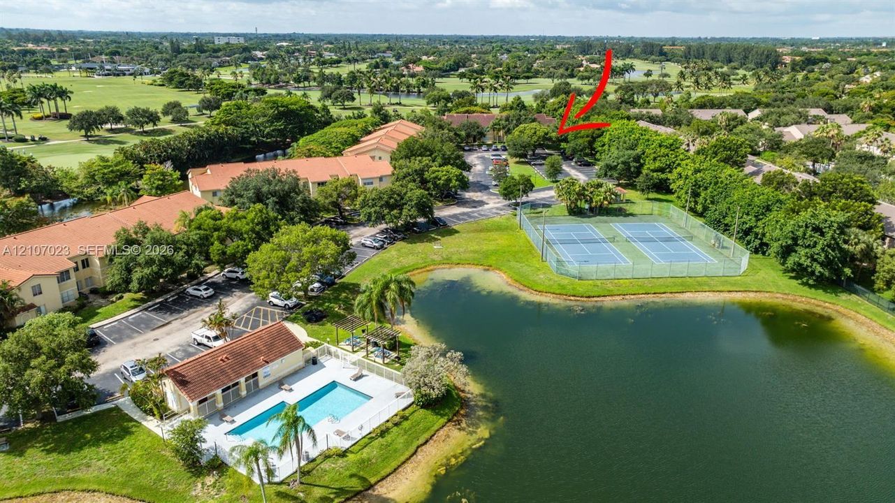 Aerial perspective of the community featuring an expansive lake, dual tennis courts, a swimming pool with pool house, and mature landscaping