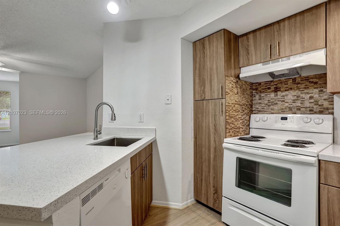 Kitchen featuring wood-finish cabinetry, stone-composite countertops, a mosaic tile backsplash, a stainless steel undermount sink with a gooseneck faucet, and white appliances