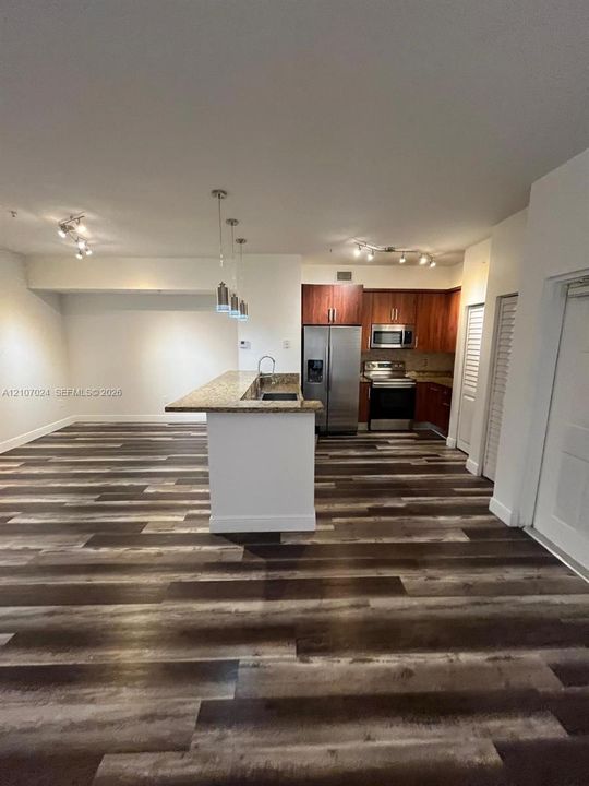 Kitchen featuring wood-finish flooring, stone-surfaced island with integrated sink, wood-tone cabinetry, stainless steel appliances, and multi-bulb track lighting