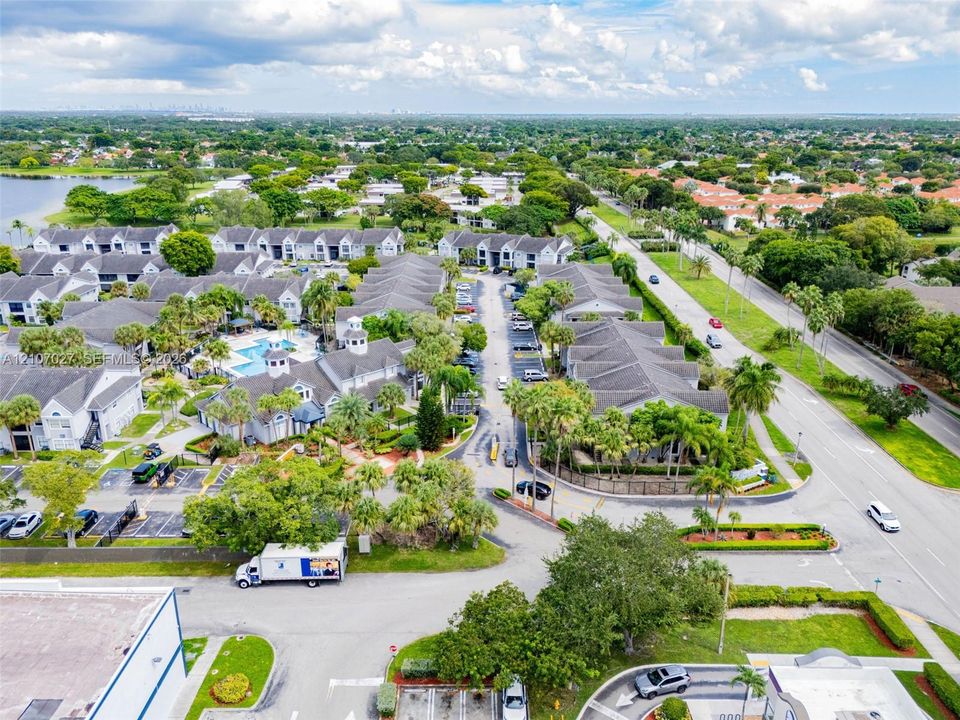 Aerial view of a residential community featuring low-rise buildings with grey shingle roofing, mature palm trees, landscaped green spaces, and a shared swimming pool