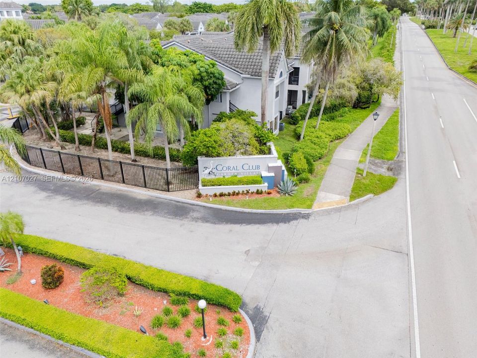 Community entrance featuring a concrete monument sign, mature palm trees, manicured hedges, and perimeter metal fencing