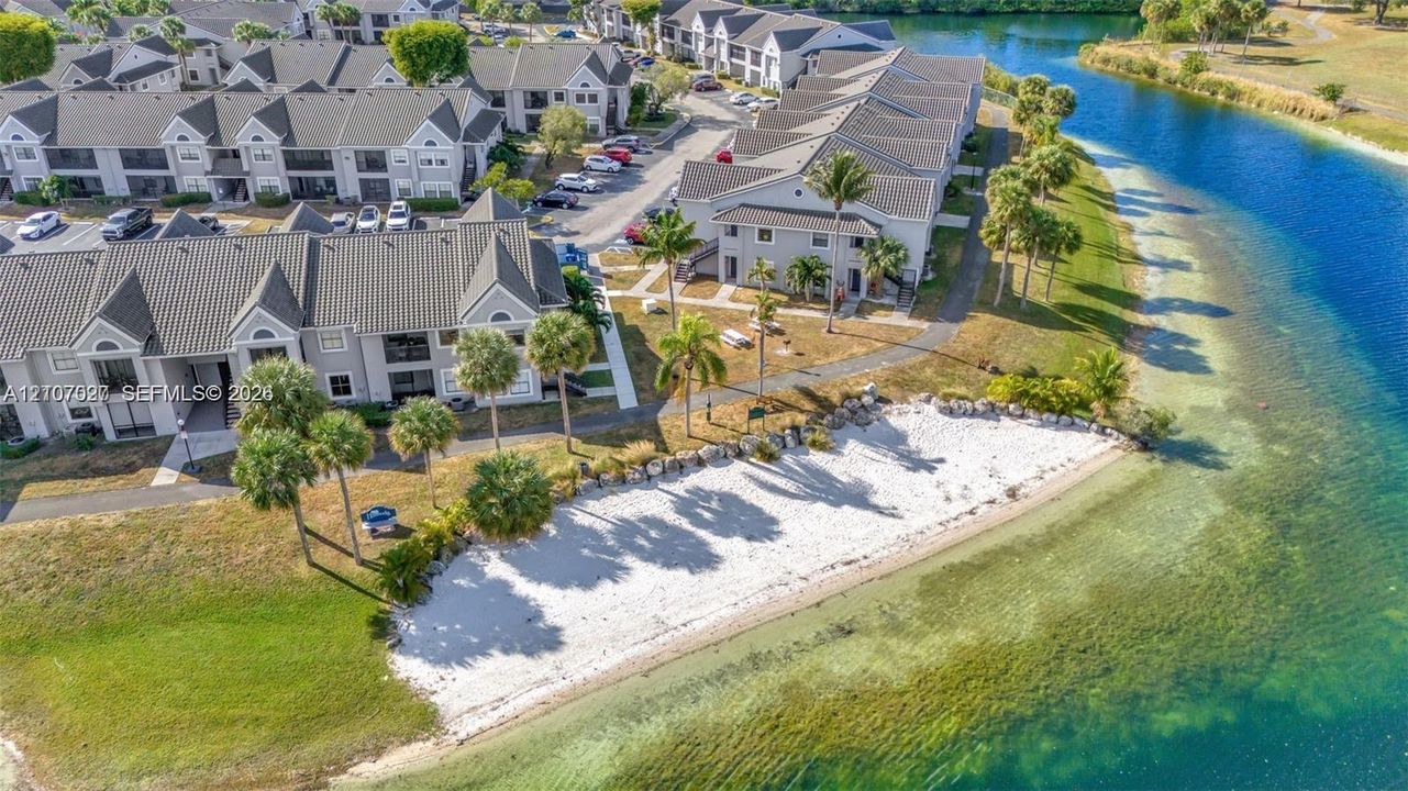 Aerial perspective of multi-unit residential structures featuring a sandy shoreline, paved walking paths, and mature palm trees situated along a waterfront