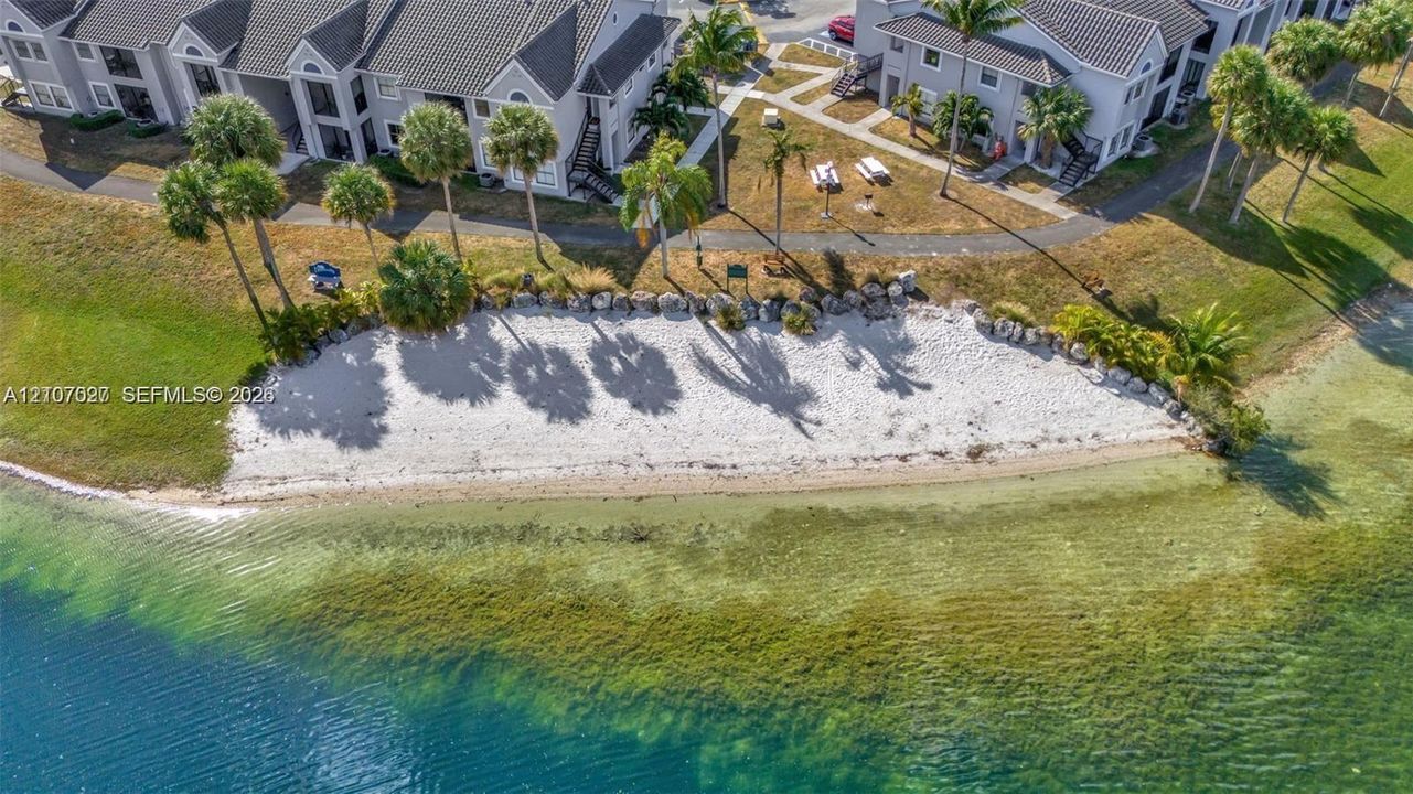 Waterfront property featuring a sandy shoreline, rock landscaping, mature palm trees, and paved walking paths