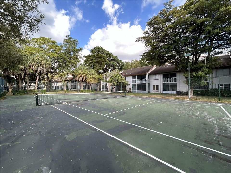 Tennis court with chain-link fencing, mature trees, and multi-level residential building exterior