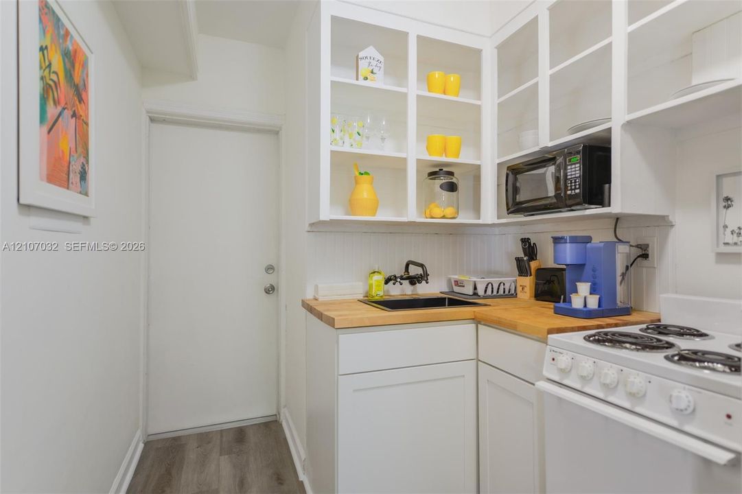 Kitchenette featuring white shaker-style cabinetry, open shelving with glass-front uppers, butcher block countertops, a black undermount sink with a matte black faucet, and wood-finish flooring