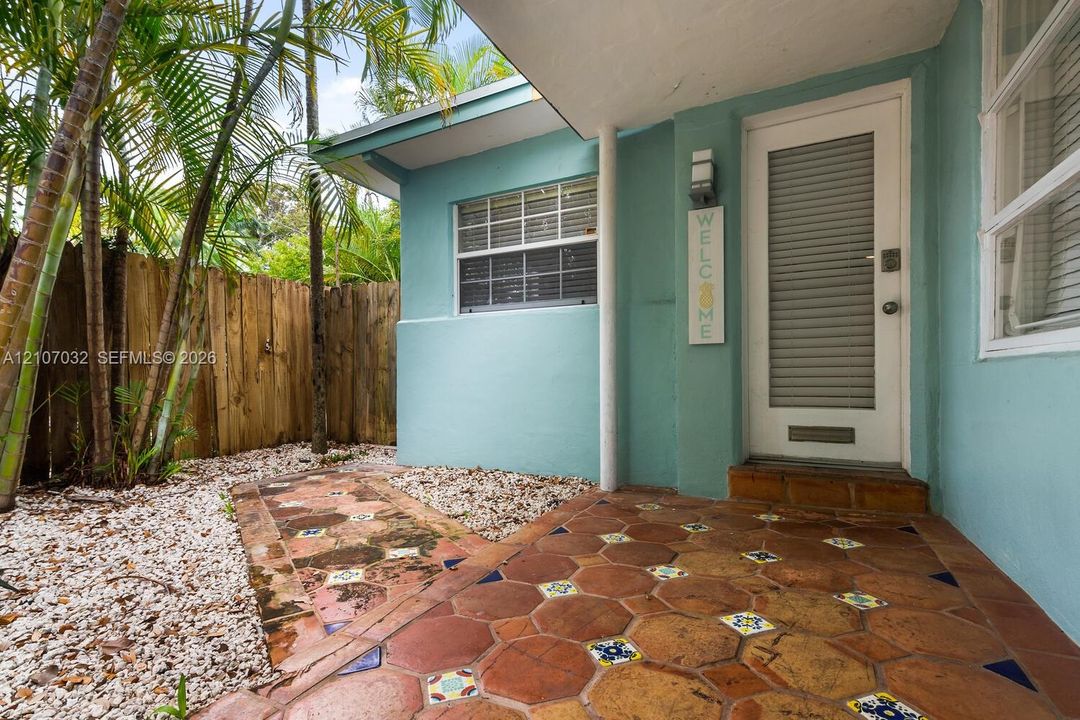 Entryway featuring a patterned terracotta tile porch, louvered glass door, smooth-finish exterior siding, stone-ground landscaping, and a natural wood privacy fence