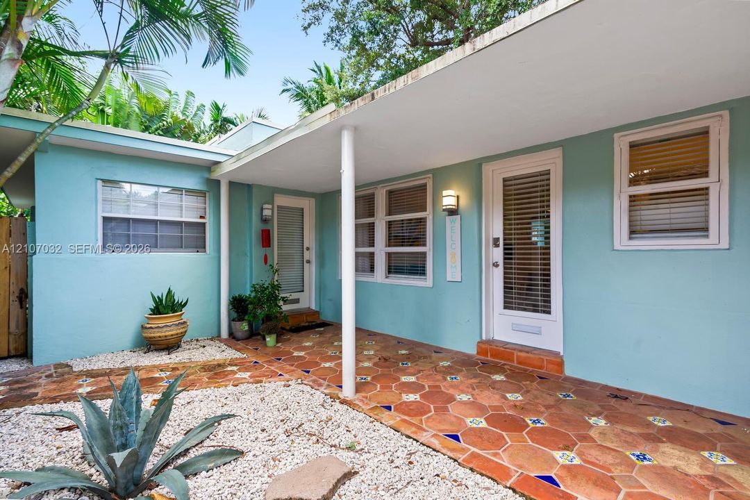 Exterior facade featuring light-colored stucco, a covered porch with patterned tile flooring, dual glass-paneled entry doors, and gravel landscaping